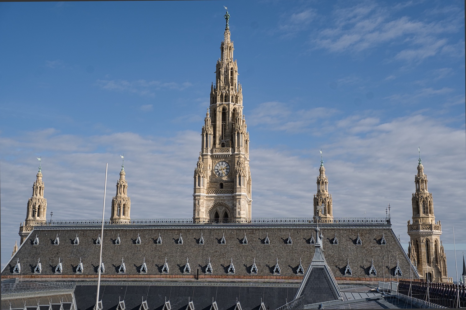 Blick vom Dach auf das Wiener Rathaus: neogotischer Bau mit zentralem Turm, Uhr und filigranen Türmchen vor blauem Himmel.