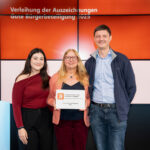 Zwei Frauen und ein Mann in Gruppenfoto-Aufstellung, die Frau in der Mitte hält eine Preisurkunde.