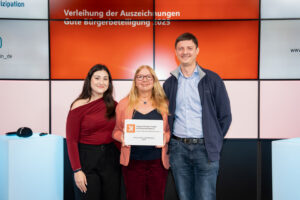 Zwei Frauen und ein Mann in Gruppenfoto-Aufstellung, die Frau in der Mitte hält eine Preisurkunde.