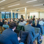 Blick über die Rückn und Schultern des Publikums auf eine Konferenzbühne.