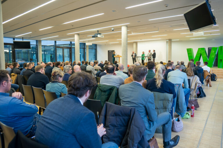 Blick über die Rückn und Schultern des Publikums auf eine Konferenzbühne.