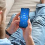 Person sitzt entspannt auf einem Sofa und hält ein Smartphone in den Händen, auf dessen Bildschirm das Logo und die Aufschrift „EUDI-Wallet“ mit EU-Sternenkreis zu sehen sind.