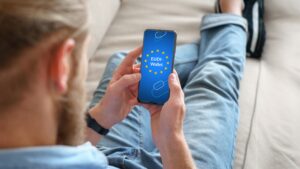 Person sitzt entspannt auf einem Sofa und hält ein Smartphone in den Händen, auf dessen Bildschirm das Logo und die Aufschrift „EUDI-Wallet“ mit EU-Sternenkreis zu sehen sind.