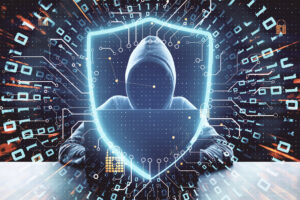 Silhouette einer gesichtslosen Person mit Kapuzenpulli die auf einer Tastatur tippt, im Bildvordergrund ist ein abstrakt dargestelltes Schutzschild zu sehen.