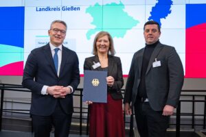 Eine Frau steht zwischen zwei Männern und hält die Förderurkunde für den Landkreis Gießen in den Händen.