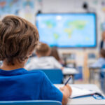 Mehrere Schüler sitzen in einem Klassenzimmer, eine Lehrerin unterrichtet sie mit Unterstützung eines Whiteboards.