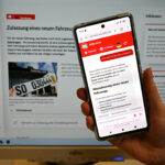 Hochgehaltenes Smartphone mit Chatbot-Screen, im Hintergrund Monitor mit Website des Kreis Soest.