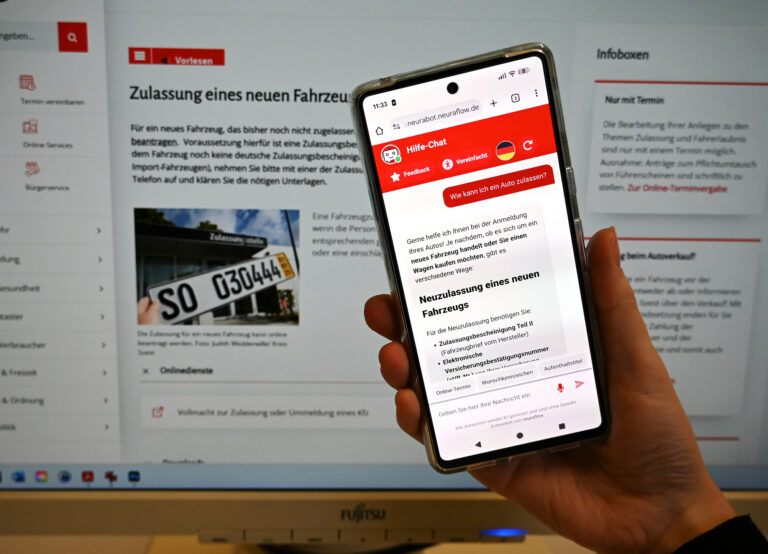 Hochgehaltenes Smartphone mit Chatbot-Screen, im Hintergrund Monitor mit Website des Kreis Soest.