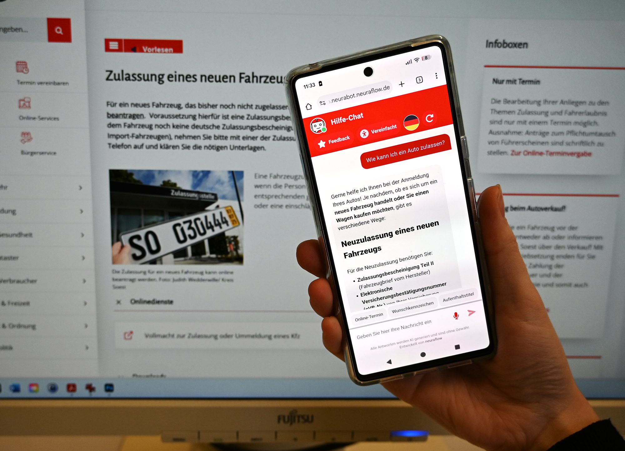 Hochgehaltenes Smartphone mit Chatbot-Screen, im Hintergrund Monitor mit Website des Kreis Soest.