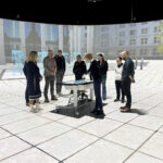 Mehrere Personen stehen an einem kleinen Tisch versammelt, im Hintergrund ist das virtuelle Modell eines Platzes in einer Stadt zu sehen.