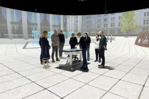 Mehrere Personen stehen an einem kleinen Tisch versammelt, im Hintergrund ist das virtuelle Modell eines Platzes in einer Stadt zu sehen.
