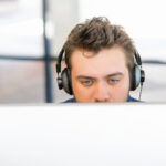 Junger Mann mit Kopfhörern in einer Büroumgebung, die untere Hälfte seines Gesichts von einem Monitor verdeckt.