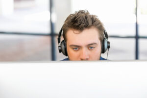 Junger Mann mit Kopfhörern in einer Büroumgebung, die untere Hälfte seines Gesichts von einem Monitor verdeckt.