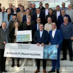 Mehrere Personen stehen zum Gruppenfoto versammelt auf einer Treppe, halten den Fördermittelbescheid und ein Banner mit der Aufschrift Echtzeit-Frühwarnsystem in den Händen.