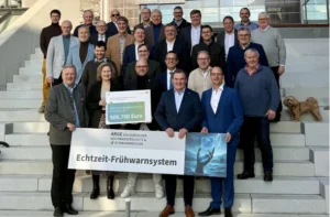 Mehrere Personen stehen zum Gruppenfoto versammelt auf einer Treppe, halten den Fördermittelbescheid und ein Banner mit der Aufschrift Echtzeit-Frühwarnsystem in den Händen.