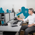 Mann im weißen Hemd sitzt vor mehreren Monitoren und zeigt auf einen davon.