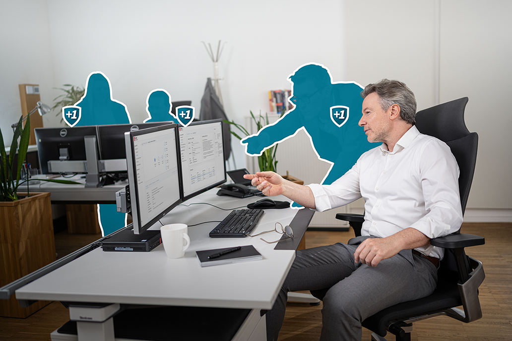 Mann im weißen Hemd sitzt vor mehreren Monitoren und zeigt auf einen davon.