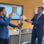 Richard Himes und Landrat Bär halten einen Taupunktsensor, der in einem Raum steht.