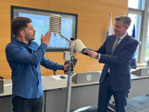 Richard Himes und Landrat Bär halten einen Taupunktsensor, der in einem Raum steht.