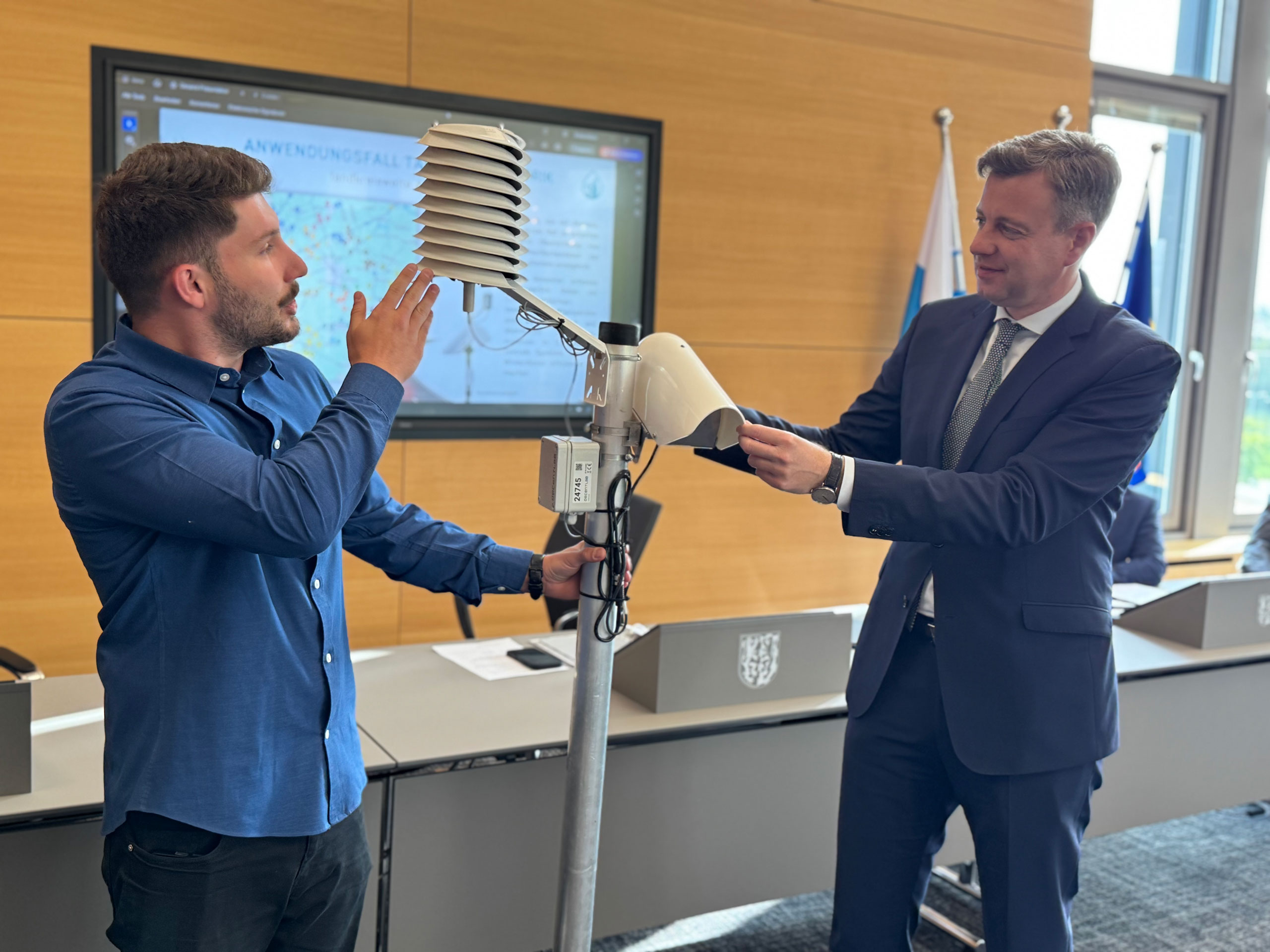 Richard Himes und Landrat Bär halten einen Taupunktsensor, der in einem Raum steht.