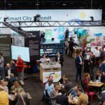 Blick auf eine Veranstaltungshalle. Im Hintergrund ist ein Messestand zu sehen, davor stehen zahlreiche kleinere Tische, an denen Personen zusammen sitzen.