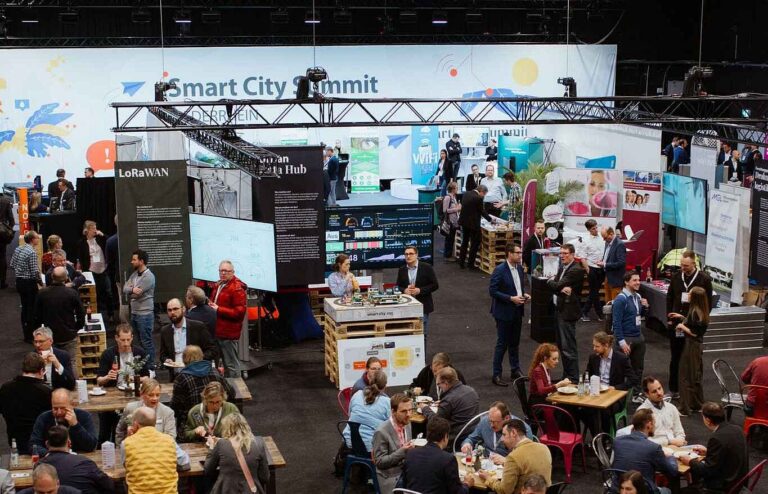 Blick auf eine Veranstaltungshalle. Im Hintergrund ist ein Messestand zu sehen, davor stehen zahlreiche kleinere Tische, an denen Personen zusammen sitzen.