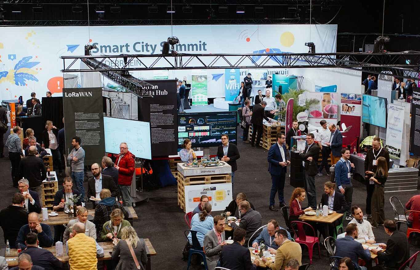 Blick auf eine Veranstaltungshalle. Im Hintergrund ist ein Messestand zu sehen, davor stehen zahlreiche kleinere Tische, an denen Personen zusammen sitzen.