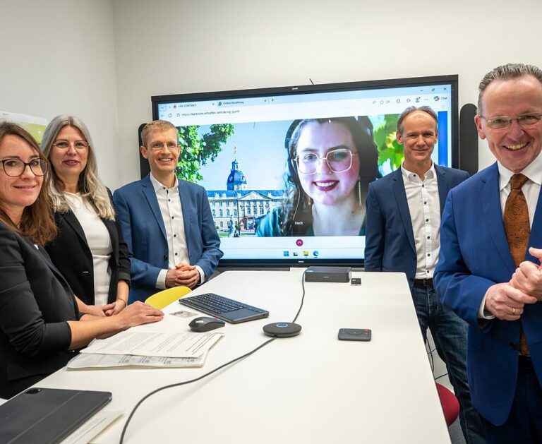 Start Videotelefonie in Karlsruhe, fünf Personen stehen am Tisch, Bildschirm im Hintergrund