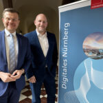 Zwei anzugtragende, lächelnde Männer neben einer Stellwand mit der Aufschrift "Digitales Nürnberg".