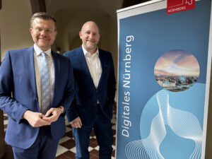 Zwei anzugtragende, lächelnde Männer neben einer Stellwand mit der Aufschrift "Digitales Nürnberg".