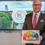 Bürgermeister Michael Gerdhenrich hält die neue Broschüre in der Hand, im Hintergrund ist auf einem Bildschirm das Video zu BE smart zu sehen.