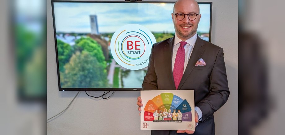 Bürgermeister Michael Gerdhenrich hält die neue Broschüre in der Hand, im Hintergrund ist auf einem Bildschirm das Video zu BE smart zu sehen.