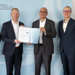 Alt-Text: Vier Männer in dunklen Anzügen stehen vor einer blauen Wand. Zwei Männer halten ein Zertifikat in die Kamera.