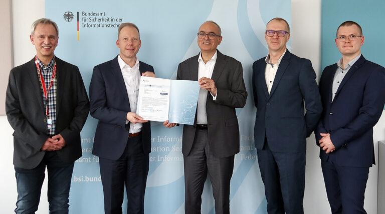 Alt-Text: Vier Männer in dunklen Anzügen stehen vor einer blauen Wand. Zwei Männer halten ein Zertifikat in die Kamera.