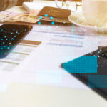 Auf einem Tisch liegen ein Smartphone und ein papierbasierter Antrag zwischen einem Laptop und anderen Utensilien auf einem Tisch.