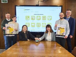 Mehrere Personen stehen vor einem Whiteboard mit einer Präsentation der Integriert-App versammelt, zwei davon drücken symbolisch den Startknopf des neuen Angebots.