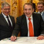 Drei offensichtlich gut gelaunte, lachende Männer in formaler Kleidung vor einem Tisch mit Dokumenten, warme Farbstimmung im Bild.