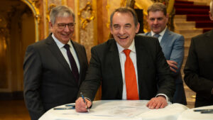 Drei offensichtlich gut gelaunte, lachende Männer in formaler Kleidung vor einem Tisch mit Dokumenten, warme Farbstimmung im Bild.