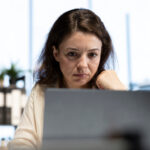 Frau in einem hellen Büro starrt mit genervtem Gesichtsausdruck auf einen Computerbildschirm