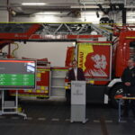 Zwei Personen stehen vor einem Feuerwehrauto, neben ihnen befindet sich ein Display, das die Oberfläche der Software rescueTablet angezeigt wird.