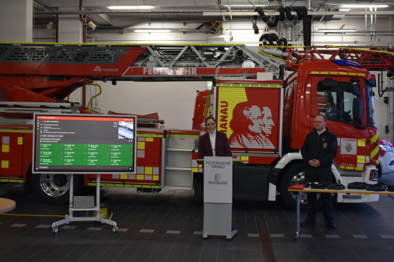 Zwei Personen stehen vor einem Feuerwehrauto, neben ihnen befindet sich ein Display, das die Oberfläche der Software rescueTablet angezeigt wird.