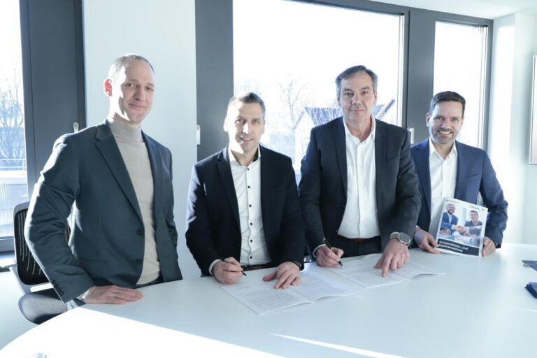 Vier Personen stehen nebeneinander an einem Tisch versammelt, zwei von ihnen unterzeichnen den Vertrag zwischen OWL-IT und regio iT.