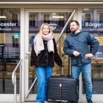 Eine Frau und ein Mann stehen mit einem zugebundenen Rollkoffer vor der Glasfront des "Bürgerservicecenters der Landeshauptstadt Potsdam".