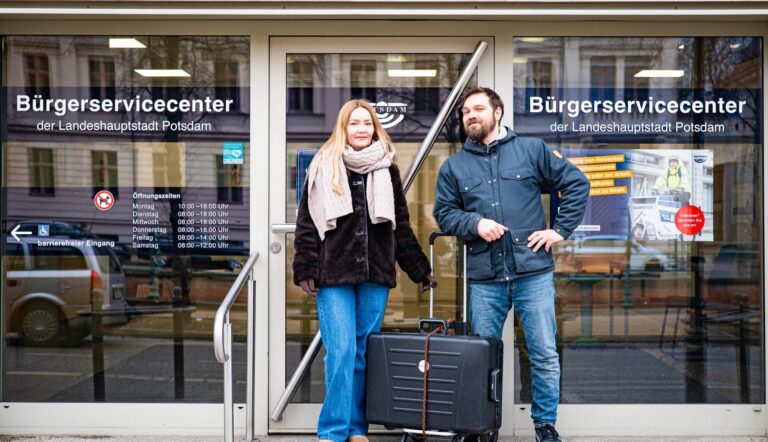 Eine Frau und ein Mann stehen mit einem zugebundenen Rollkoffer vor der Glasfront des "Bürgerservicecenters der Landeshauptstadt Potsdam".
