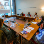 Mehrere Personen sitzen an einem U-förmigen Besprechungstisch, eine Person ist virtuell via Laptop zugeschaltet.