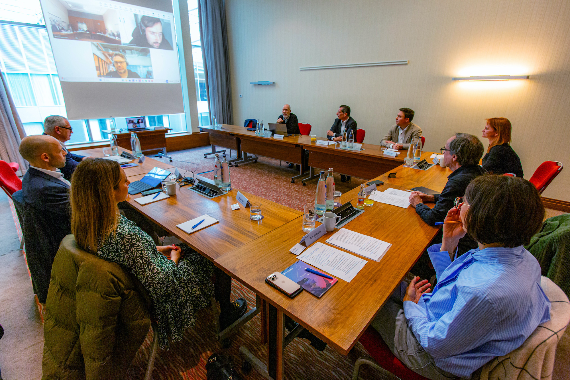 Mehrere Personen sitzen an einem U-förmigen Besprechungstisch, eine Person ist virtuell via Laptop zugeschaltet.