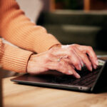 In Wollpulloverärmeln steckende Hände tippen an einem Laptop, der auf einem niedrigen Couchtisch steht. Die ganze s´Szene ist warm beleuchtet.