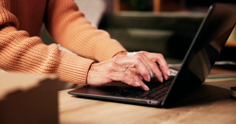 In Wollpulloverärmeln steckende Hände tippen an einem Laptop, der auf einem niedrigen Couchtisch steht. Die ganze s´Szene ist warm beleuchtet.