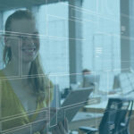 Eine lächelnde Frau hält ein Tablet in der Hand, im Hintergrund ist ein Großraumbüro zu sehen.