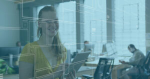 Eine lächelnde Frau hält ein Tablet in der Hand, im Hintergrund ist ein Großraumbüro zu sehen.